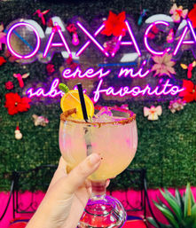 Hand holding a chili-rimmed margarita with orange slice and straw in front of a pink neon Spanish sign on a green floral wall