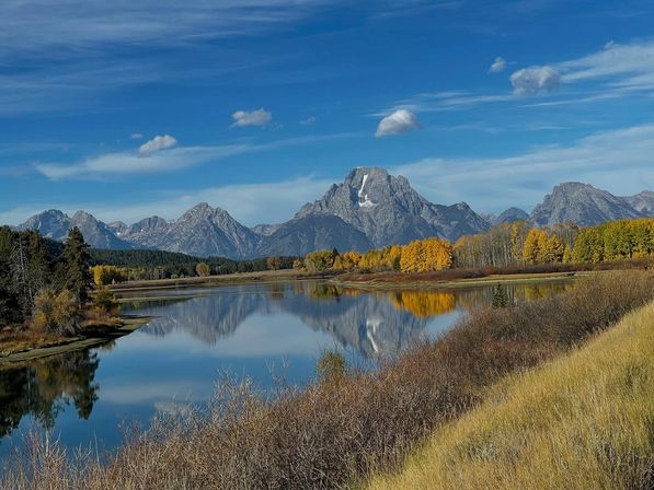 Autumn mountain landscape with snow-dusted jagged peaks mirrored in a calm river, bright golden aspen trees along the shore and a clear blue sky.