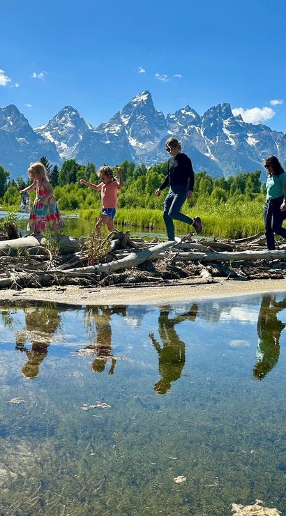 Group of four people balancing on driftwood along a calm river, their reflections in the water, grassy wetlands and snow-capped Grand Teton Range in Grand Teton National Park, Wyoming under a bright blue sky.
