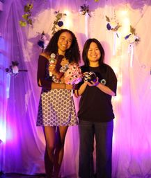 Two smiling women pose on a purple-pink lit floral backdrop at an indoor event; one holds a microphone and pink bouquet, the other holds a small black prop, with hanging flowers and fairy lights behind them.