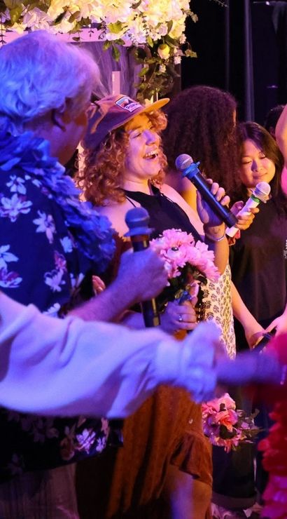 Mixed-age group singing and cheering on an indoor stage at a lively karaoke night, purple lighting, microphones, leis and flowers