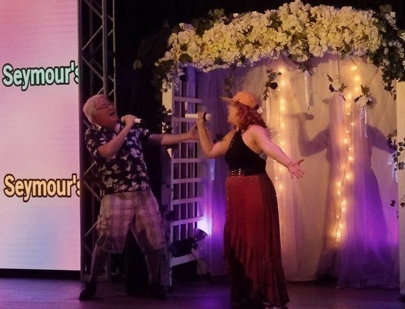 Two singers performing a lively duet with handheld microphones on an indoor stage beneath a flower-covered arch lit by string lights and purple uplighting.