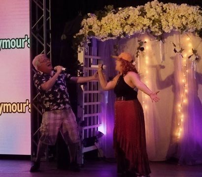 Two singers performing a lively duet with handheld microphones on an indoor stage beneath a flower-covered arch lit by string lights and purple uplighting.