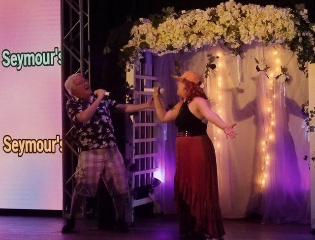 Two singers performing a lively duet with handheld microphones on an indoor stage beneath a flower-covered arch lit by string lights and purple uplighting.