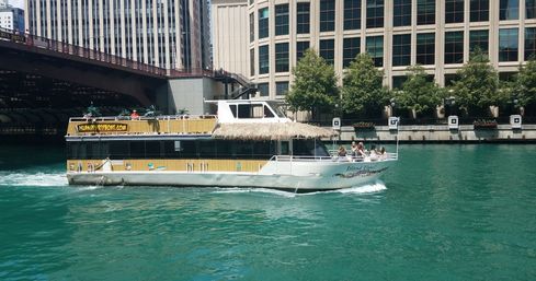 Thatched-roof tour boat cruising bright turquoise downtown riverfront under a city bridge, passengers relaxing on the open-air deck by sunlit high-rise buildings