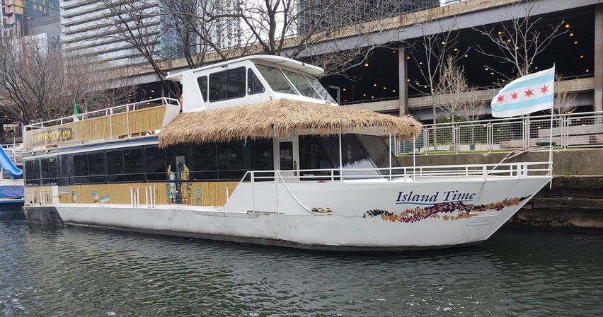 White two-story river tour boat with a thatched tiki-style roof docked along an urban riverwalk, decorative floral hull artwork and city buildings in the background.