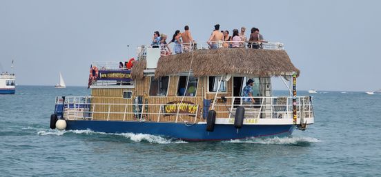 Thatched-roof party boat cruising coastal waters with people on the sunlit upper deck, sailboats dotting the horizon for a lively summer boat tour vibe.