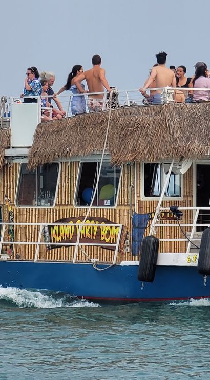 Thatched-roof party boat cruising coastal waters with people on the sunlit upper deck, sailboats dotting the horizon for a lively summer boat tour vibe.