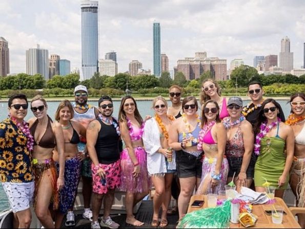 Group of friends in colorful leis, swimsuits and hula skirts enjoying a sunny boat party on the water with an urban skyline and waterfront park in the background.