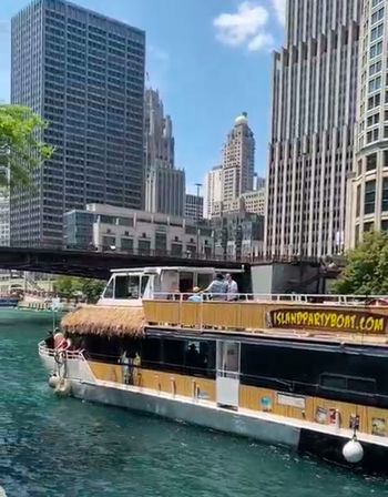 Tiki-style yellow party boat cruising a turquoise downtown river, passing under a bridge with people on deck and tall skyscrapers against a sunny blue sky