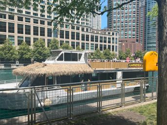 Tiki-style party boat docked at a downtown riverwalk with passengers on the upper deck, tall office buildings and leafy trees along the waterfront, and a bright yellow life ring station visible.
