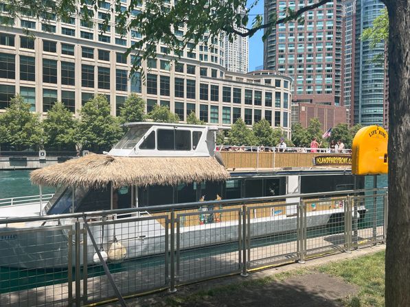 Tiki-style party boat docked at a downtown riverwalk with passengers on the upper deck, tall office buildings and leafy trees along the waterfront, and a bright yellow life ring station visible.