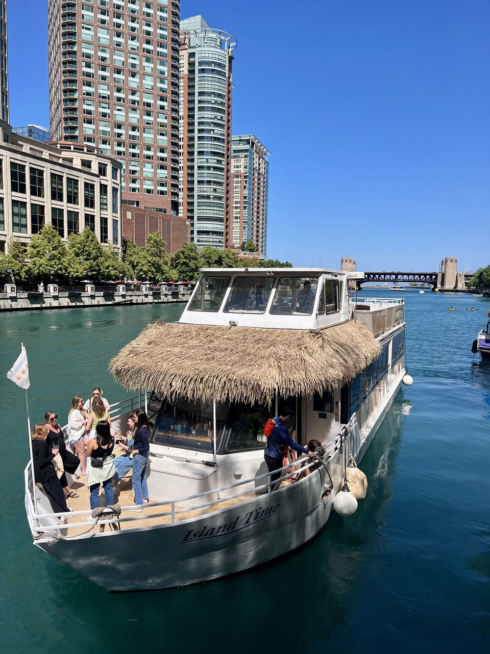 Thatched roof leisure boat on the Chicago River with people on deck and downtown skyscrapers under a bright blue sky