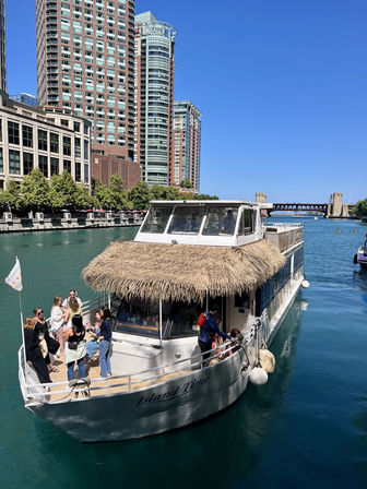 Thatched roof leisure boat on the Chicago River with people on deck and downtown skyscrapers under a bright blue sky