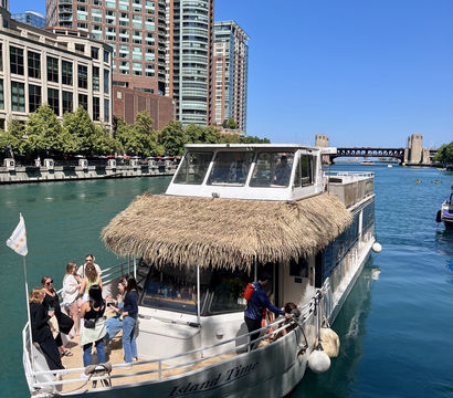 Thatched roof leisure boat on the Chicago River with people on deck and downtown skyscrapers under a bright blue sky