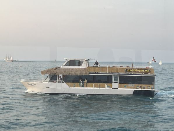 Tiki-roofed double-decker party boat cruising calm coastal waters with an American flag and sailboats on the horizon under a pale evening sky