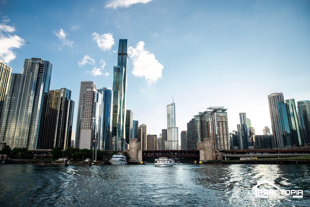 Chicago River skyline with glass skyscrapers, a low bridge and a tour boat on rippling water under a bright blue sky