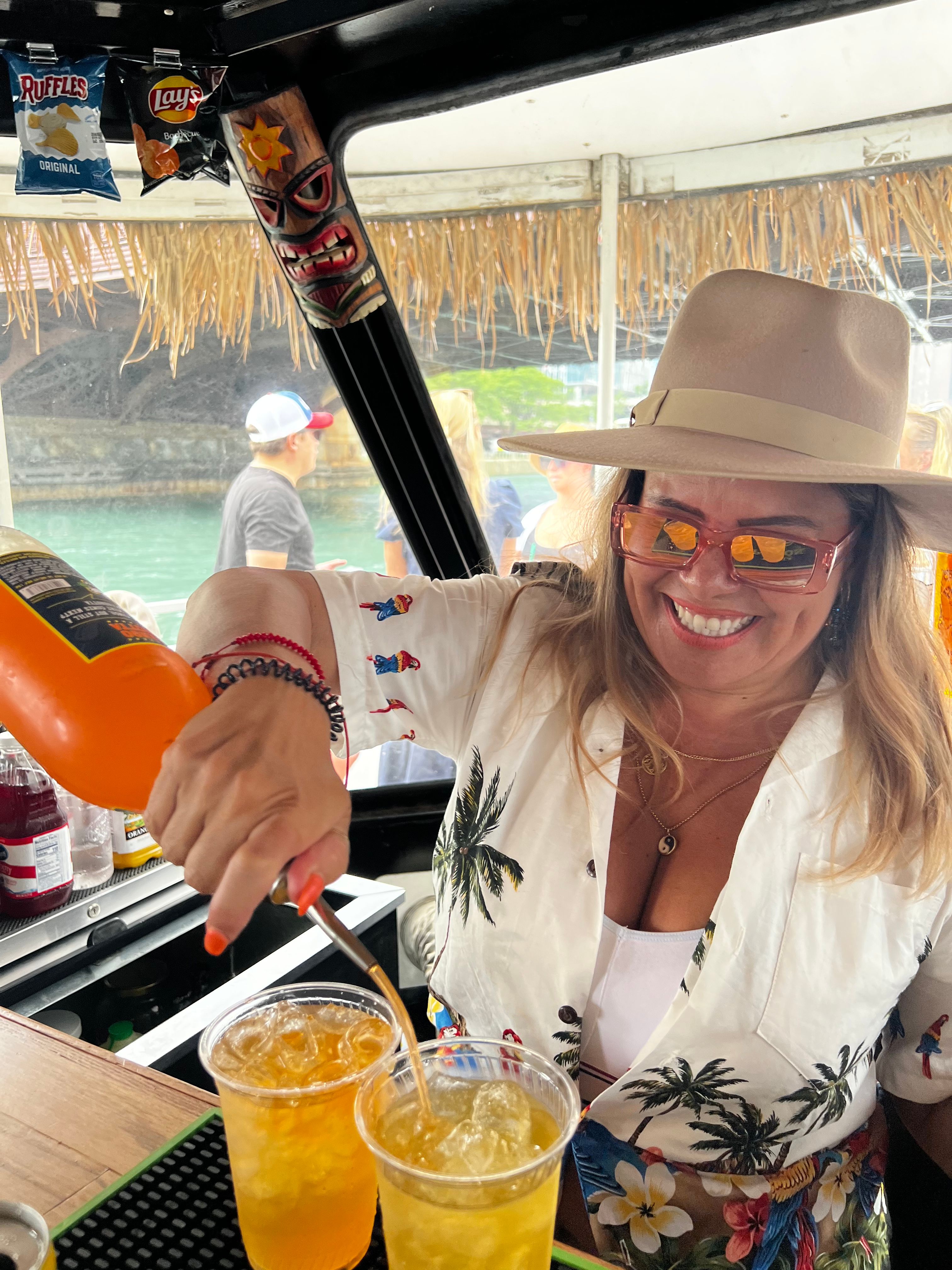 Smiling bartender in a sun hat and sunglasses pouring tropical cocktails into plastic cups at a tiki-decor boat bar on turquoise waterfront