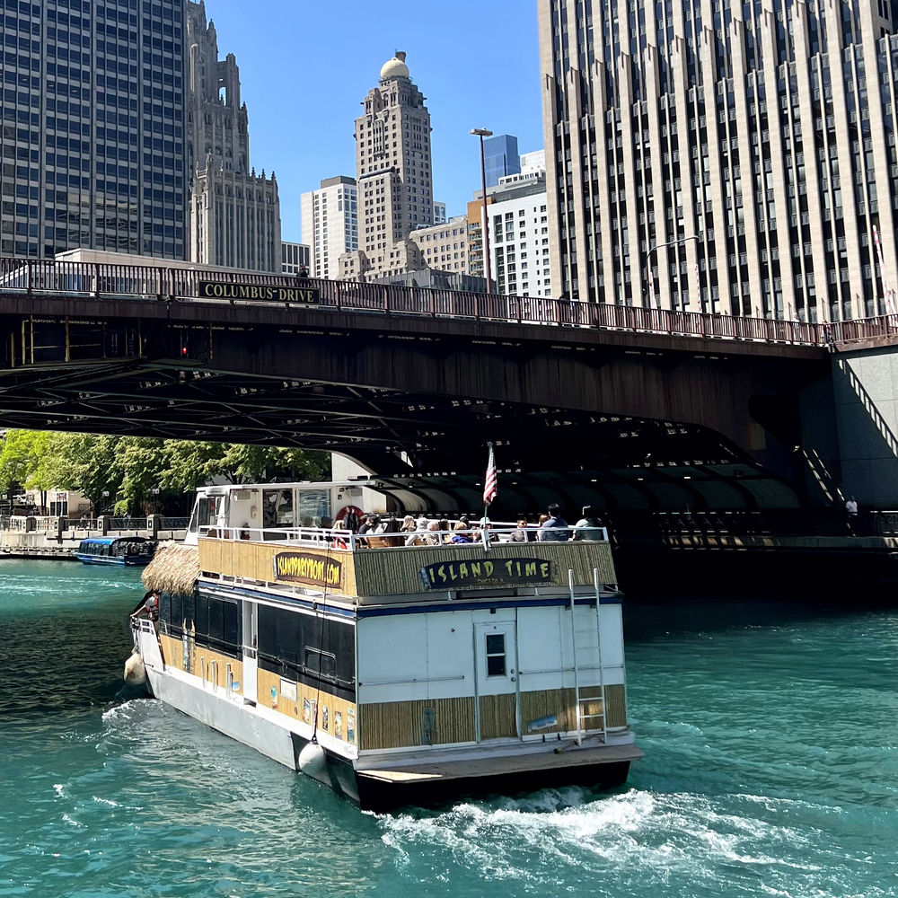 Tour boat cruising the turquoise Chicago River beneath the Columbus Drive bridge with downtown Chicago skyscrapers on a sunny day