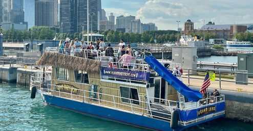 Tiki-themed party boat docked at an urban lakefront with a rooftop crowd, bright blue water slide, American flag, and downtown skyline in the background on a sunny day.