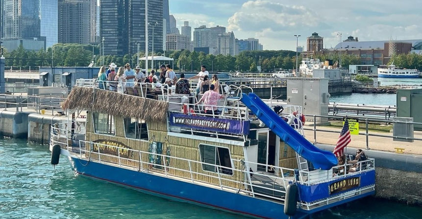 Tiki-themed party boat docked at an urban lakefront with a rooftop crowd, bright blue water slide, American flag, and downtown skyline in the background on a sunny day.