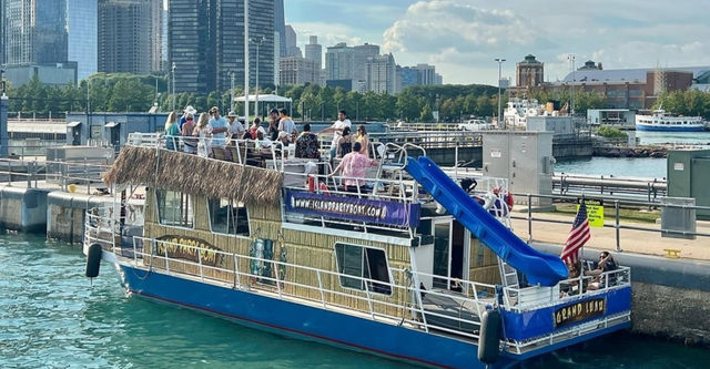 Tiki-themed party boat docked at an urban lakefront with a rooftop crowd, bright blue water slide, American flag, and downtown skyline in the background on a sunny day.