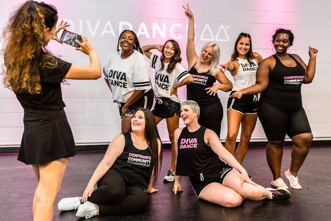 Eight women in athletic wear laughing and striking playful poses for a photo inside a bright indoor dance studio after a group dance class.