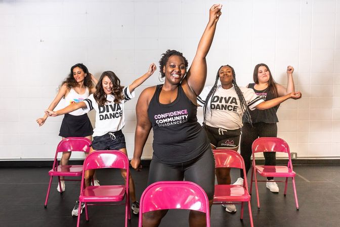 Five women in athletic wear perform a synchronized chair-dance routine in a bright studio, smiling and striking energetic arm-up poses behind pink folding chairs.