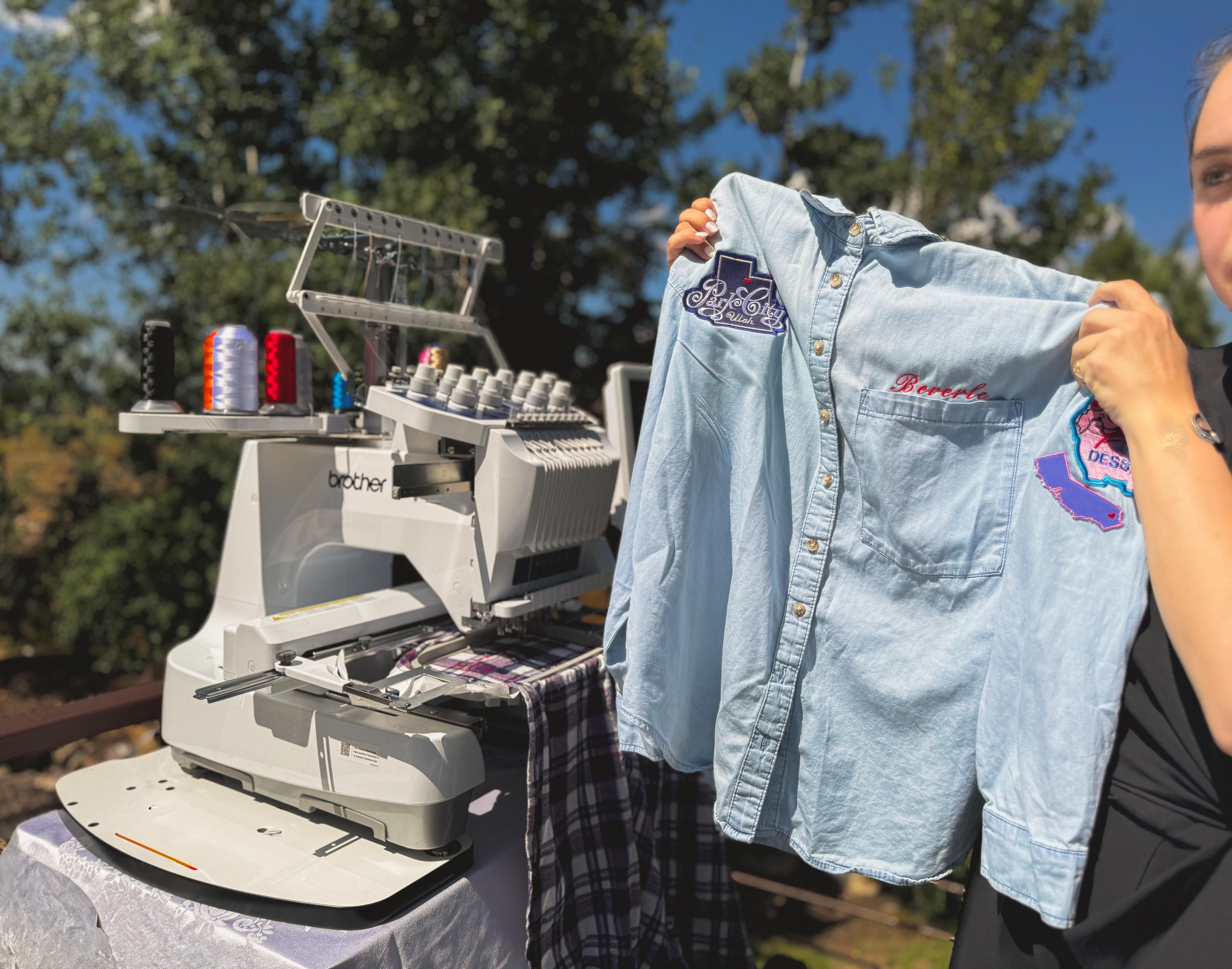 Outdoor embroidery setup with a Brother multi-needle machine and colorful thread spools, a light denim shirt held up to display custom embroidered patches and a stitched name under bright sunlight