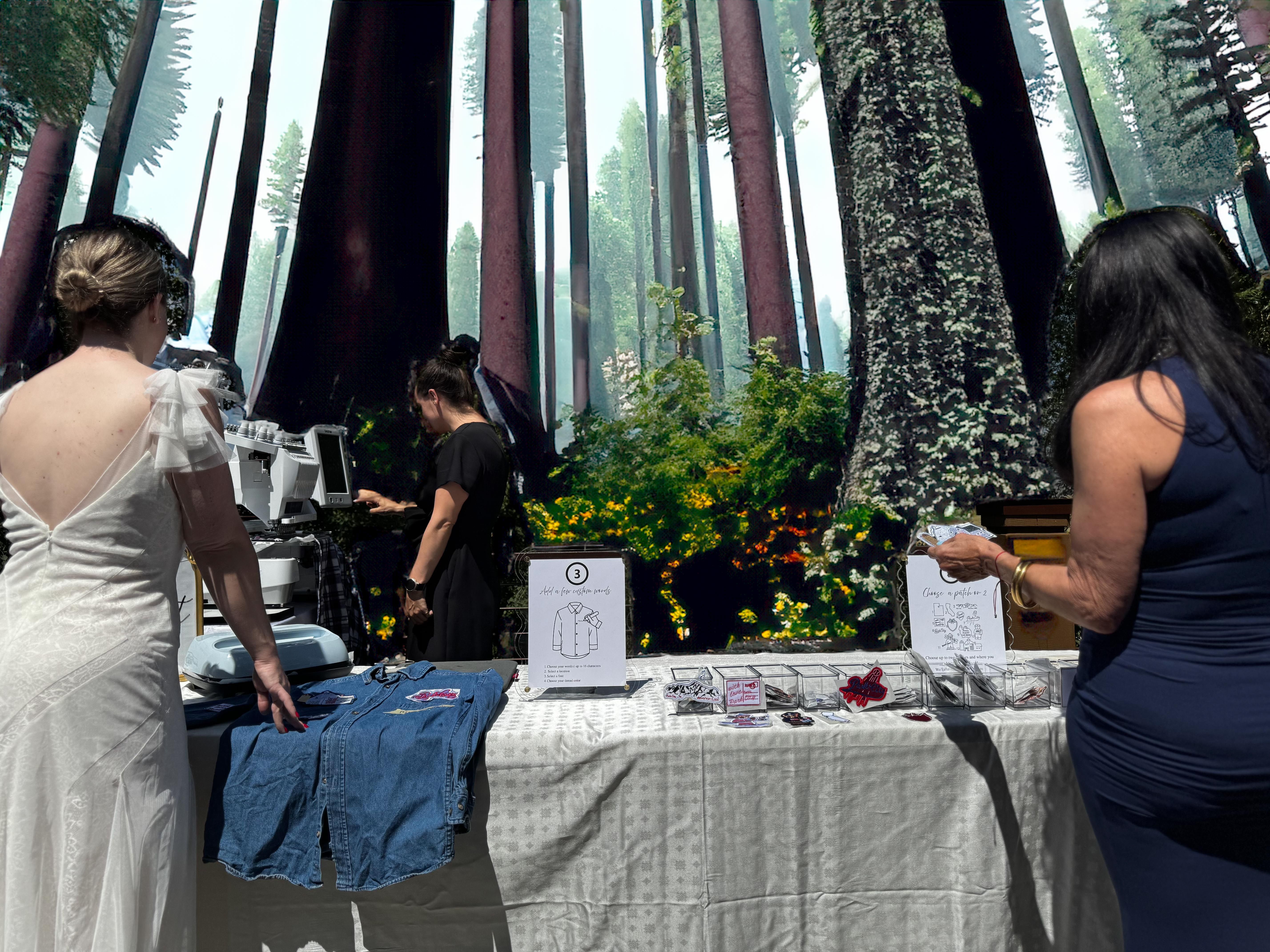 Three women at a DIY craft station adding patches to a denim jacket in front of a giant redwood forest mural