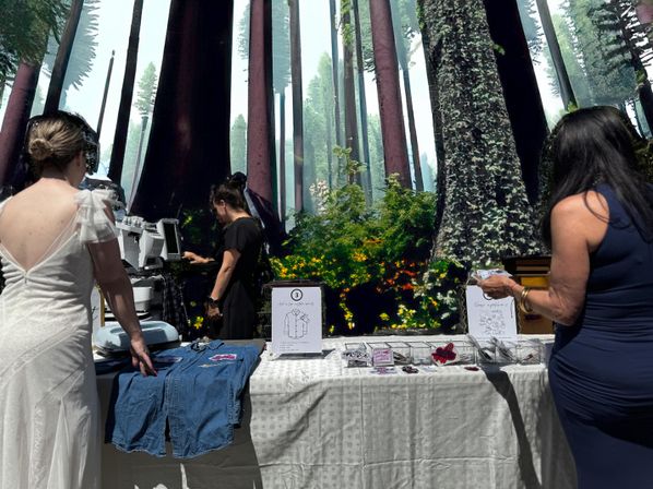 Three women at a DIY craft station adding patches to a denim jacket in front of a giant redwood forest mural
