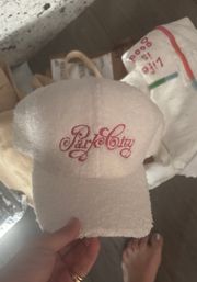 Hand holding a small plush white terry-cloth hotel-style visor with pink cursive embroidery, paper bags and a bare foot on a wood floor in the background.