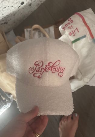 Hand holding a small plush white terry-cloth hotel-style visor with pink cursive embroidery, paper bags and a bare foot on a wood floor in the background.