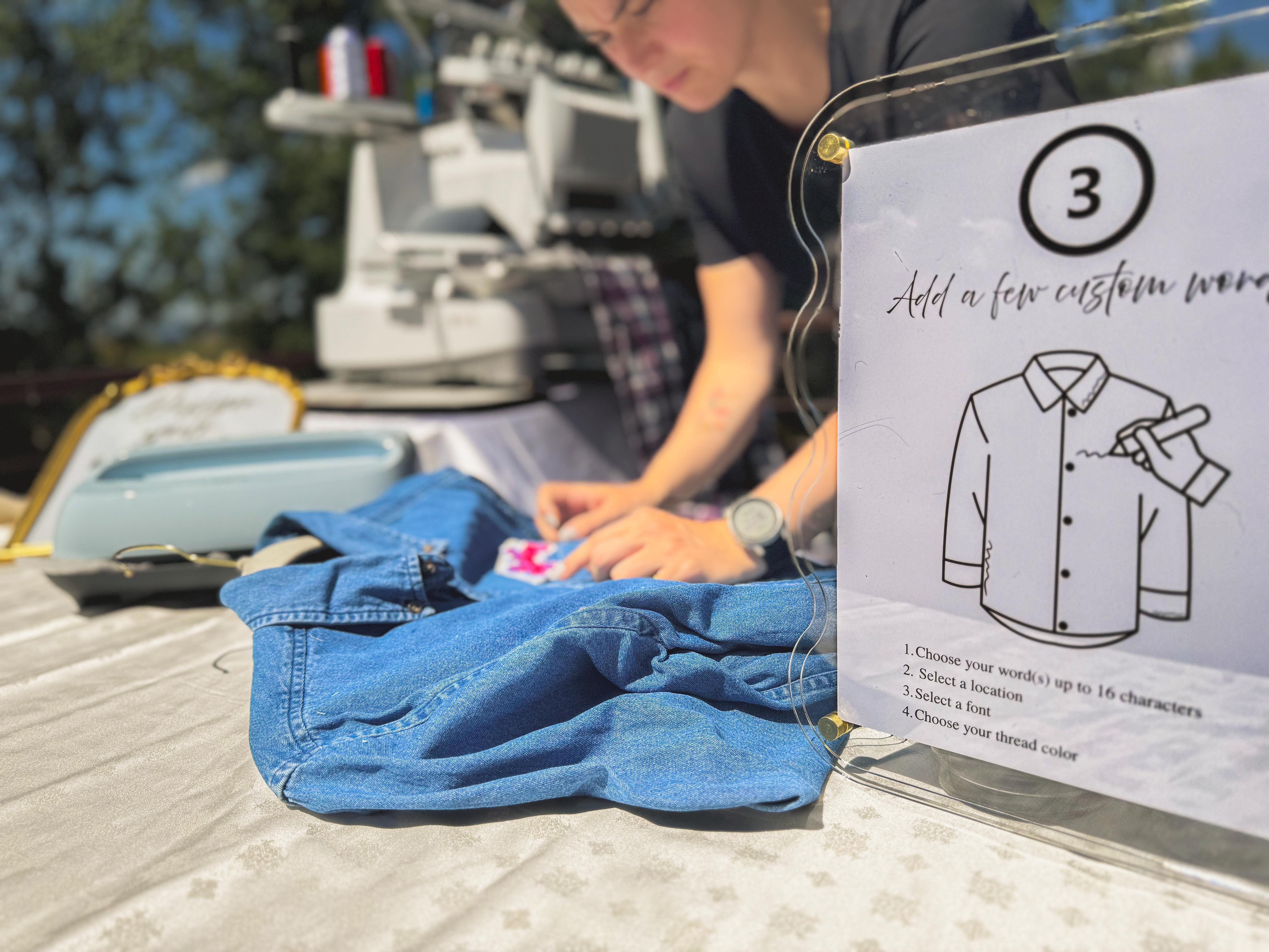 Denim jacket on a vendor table at an outdoor market with an embroidery machine adding a colorful patch and a nearby sign showing custom shirt personalization steps.