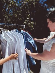 Woman browsing white shirts and denim jackets on an outdoor clothing rack at a sunny pop-up market