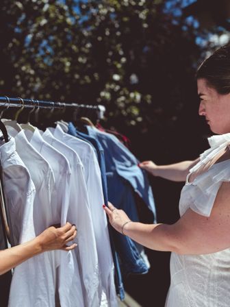 Woman browsing white shirts and denim jackets on an outdoor clothing rack at a sunny pop-up market