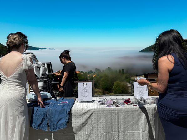 Hilltop outdoor vendor table with denim patches and small crafts, three women arranging items at a scenic overlook while coastal fog fills the valley below under a clear blue sky.