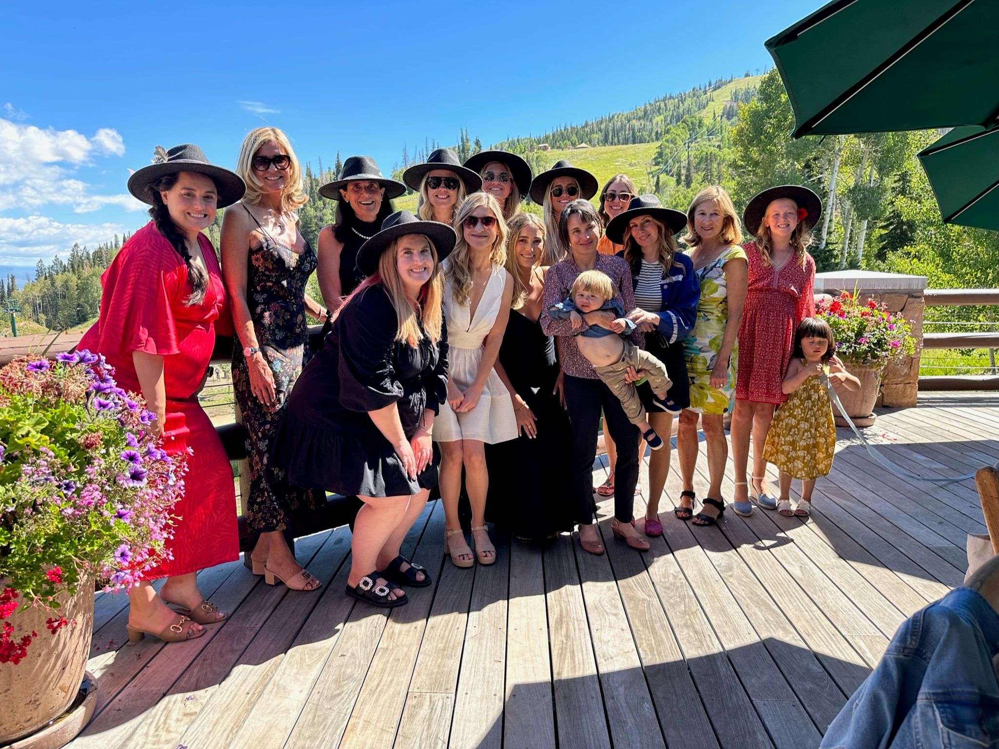 Smiling group of women and children on a sunny mountain deck, many wearing wide-brim hats and summer dresses, colorful flower pots and a forested hillside under a bright blue sky.