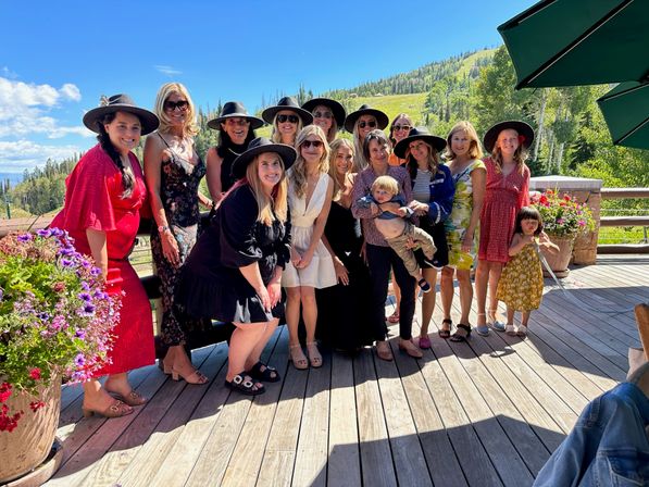 Smiling group of women and children on a sunny mountain deck, many wearing wide-brim hats and summer dresses, colorful flower pots and a forested hillside under a bright blue sky.