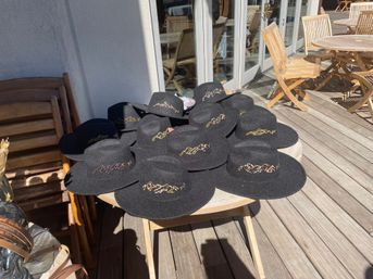 Sunlit wooden deck patio with a round table stacked with black wide-brim felt hats adorned with gold embroidered mountain motifs.