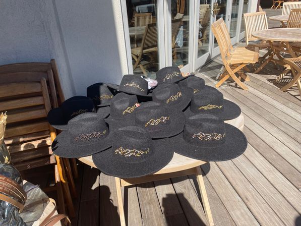 Sunlit wooden deck patio with a round table stacked with black wide-brim felt hats adorned with gold embroidered mountain motifs.