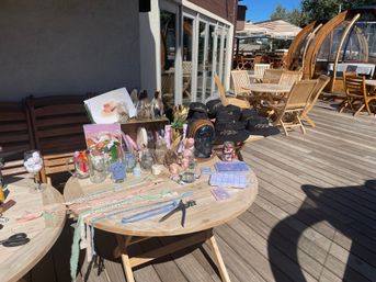 Sunny outdoor wooden deck patio with round tables showcasing a craft display of glass jars, pastel ribbons, small trinkets, notebooks and a stack of black embroidered hats beside wooden dining chairs and domed outdoor dining pods.