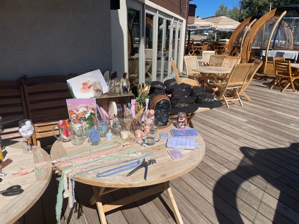 Sunny outdoor wooden deck patio with round tables showcasing a craft display of glass jars, pastel ribbons, small trinkets, notebooks and a stack of black embroidered hats beside wooden dining chairs and domed outdoor dining pods.