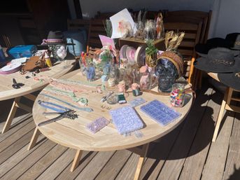 Sunlit wooden patio table on a deck piled with DIY craft supplies and decor — jars of ribbons, buttons and beads, scissors and hole punches, floral stems, mason jars, a decorative skull and black felt hats.