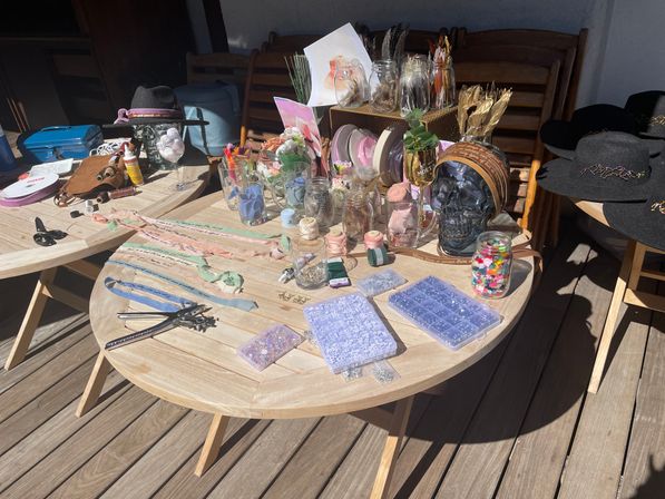 Sunlit wooden patio table on a deck piled with DIY craft supplies and decor — jars of ribbons, buttons and beads, scissors and hole punches, floral stems, mason jars, a decorative skull and black felt hats.
