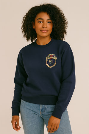 Studio portrait of a young woman with curly hair wearing a navy crewneck sweatshirt with an embroidered gold crest patch reading "MH" and light-wash jeans against a neutral beige background.
