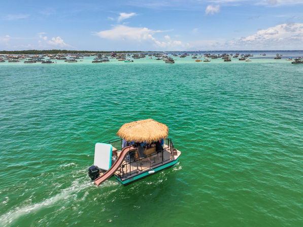 Pontoon boat with a thatched tiki roof and slide cruising on clear turquoise coastal waters, with dozens of anchored boats in the shallow bay under a sunny blue sky.