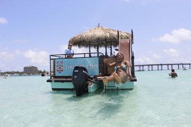 Smiling woman in a bikini slides from a thatched-roof floating tiki bar into clear turquoise shallow water on a sunny day, with staff on the boat and a distant bridge in the background.
