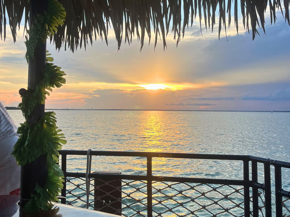 Tiki-roofed waterfront deck with lei-wrapped post and metal railing overlooking a golden sunset reflecting on calm bay water