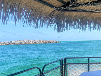 Relaxing view from a palapa‑shaded deck over turquoise ocean with metal railing and rope netting, rocky breakwater and small channel marker under a clear blue sky